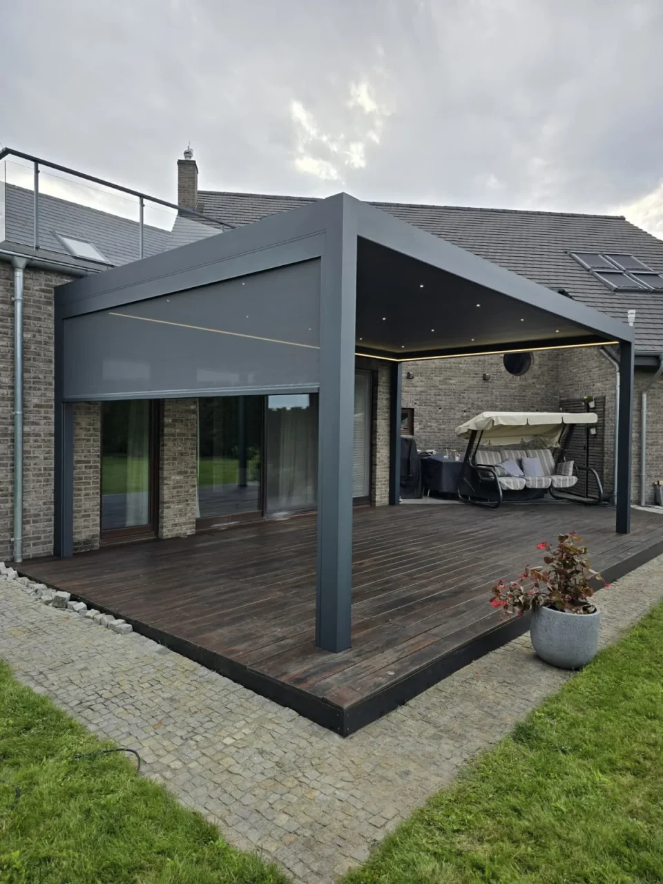 Pergola z oświetleniem i częściowo opuszczoną osłoną przy ceglanym domu