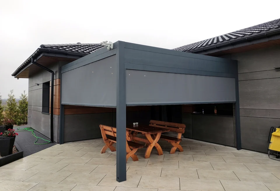 Grafitowa pergola z opuszczoną osłoną nad tarasem z drewnianym stołem