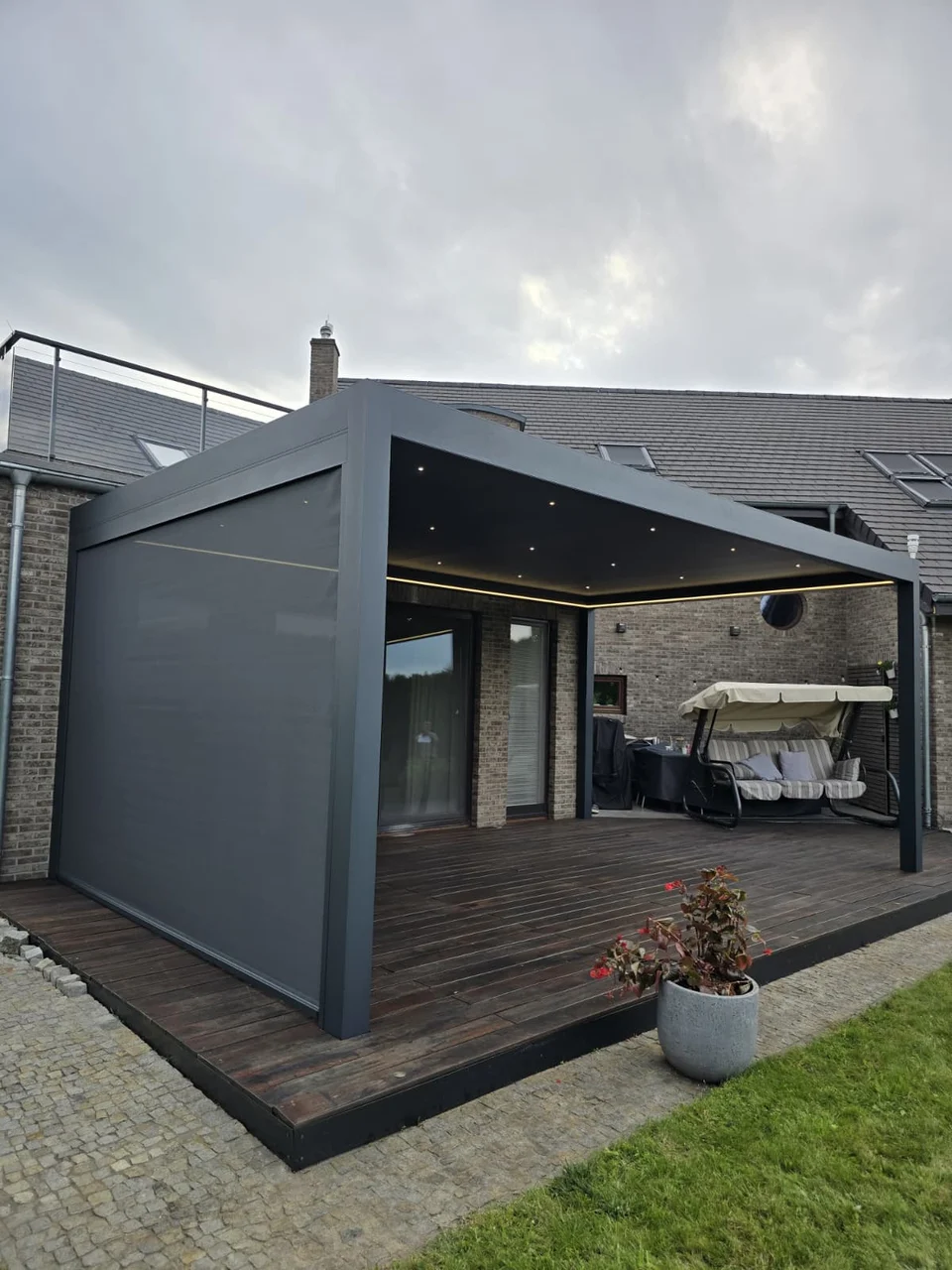 Pergola z oświetleniem i opuszczoną osłoną nad drewnianym tarasem