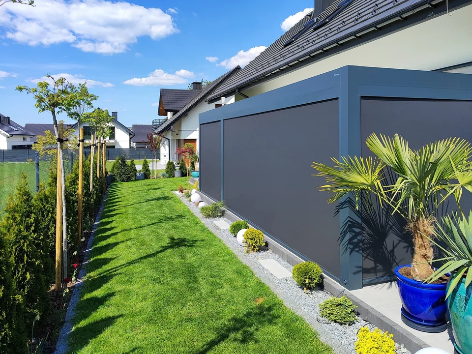 Pergola z opuszczoną osłoną boczną przy trawniku i palmach w donicach